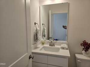 A small bathroom with a white vanity, silver faucet, and a rectangular mirror. A hand towel hangs on the wall, soap and folded towels are on the counter, and red flowers sit on the toilet tank. A door is partially open.