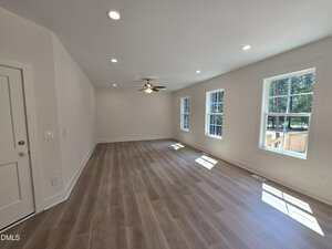 A spacious, empty room with light-colored wood flooring, white walls, recessed ceiling lights, a ceiling fan, and several large windows letting in natural light.
