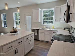 A modern kitchen with white cabinets, stainless steel appliances, a central island with fruit decor, large windows, and light-colored wood flooring, all well-lit by natural light.