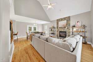 A spacious living room with a light gray sectional sofa, stone fireplace, wooden shelves, and hardwood floors. An open dining area with a table and chairs is visible in the background. Walls are painted a light gray.