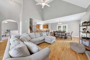 A spacious open-concept living and dining area with light gray walls, a large sectional sofa, wooden floors, a dining table with chairs, and access to the kitchen; ceiling fan and natural light from multiple windows.