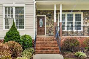 A brick staircase leads to a brown front door with an oval window, flanked by white trim, stone siding, and a metal railing. Bushes and shrubs line the front of the house. The address "2122" is visible.
