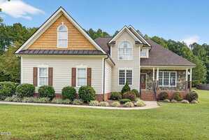 A two-story suburban house at 2122 Olde Brassfield Lane in Franklinton with beige siding, brown accents, and brick trim. The home features large windows, a covered front porch, and is surrounded by a manicured lawn and shrubbery.