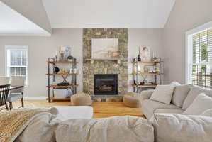 A living room with light-colored sofas, a stone fireplace centered on the back wall, two shelving units with decor on either side, and two windows letting in natural light. Wood flooring and neutral walls complete the space.