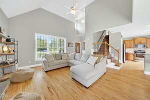 A spacious living room with light hardwood floors, a beige sectional sofa, two bean bags, a large window with blinds, open shelving, and a view of a staircase leading to an upper floor and a kitchen area.