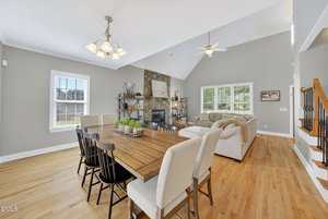 A spacious open-plan living and dining area with light wood floors, a wooden dining table and mixed chairs, a beige sectional sofa, a stone fireplace, and large windows letting in natural light.