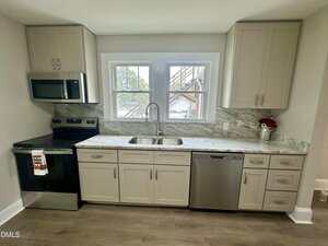 A modern kitchen with light-colored cabinets, marble-patterned countertops, stainless steel appliances including a stove, microwave, and dishwasher, and a large window above a double sink.