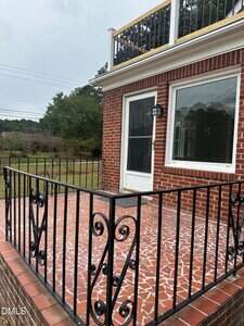 A red brick house with a white-framed glass door leads to a small patio with a red and white tile floor. The patio is enclosed by a black metal railing and overlooks a grassy yard with trees in the background.