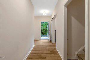 A hallway with light-colored walls and wood flooring leads to an open door revealing a view of trees outside. There are stairs on the right and a sliding barn door along the hallway.