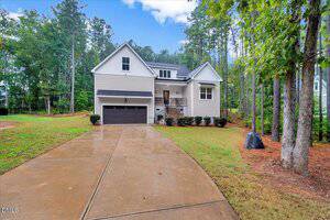 A two-story beige house with a black garage door, front porch, and gabled roof sits at the end of a wide, wet concrete driveway, surrounded by grass, pine trees, and wooded areas.
