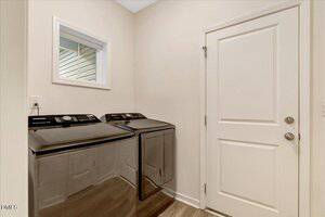 A laundry room with a front-loading washer and dryer side by side, a small window above the appliances, beige walls, wood-look flooring, and a white door to the right.