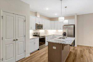A modern kitchen with white cabinets, stainless steel appliances, a marble-patterned island with a sink, light wood flooring, and pendant lighting over the island.