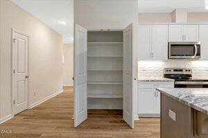 A kitchen with an open pantry closet showing four shelves. To the right are white cabinets, a stainless steel microwave, and a stove. The floor is light wood, and walls are painted beige.