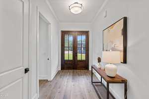 A bright entryway with light-colored walls, wood flooring, and a dark wooden front door with glass panels. A table along the right wall displays a lamp and abstract artwork hangs above it.
