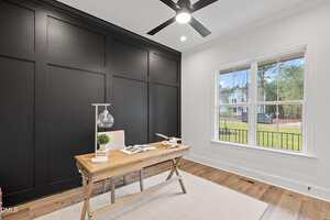 A modern home office with a wooden desk, lamp, plant, and books, set against a black accent wall. A large window lets in natural light and shows a view of houses and trees outside. The floor is light wood with a white rug.