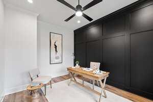 A modern home office features a wooden desk with a chair, a small round side table with a chair, a large abstract art piece on a white wall, a black accent wall, a ceiling fan, and light wood flooring.