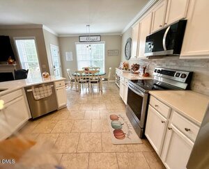 A bright kitchen with white cabinets, stainless steel appliances, beige tiled floor, and a dining table with six chairs near large windows. A decorative "Blessed" sign hangs above the table.