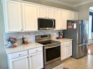 A modern kitchen with white cabinets, stainless steel appliances, a microwave above an electric stove, a refrigerator on the right, and small kitchen items on the countertops. The backsplash features light-colored tiles.