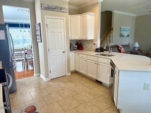 Kitchen with white cabinets and countertops, stainless steel appliances, beige tile flooring, and a pantry door. A dining area with a table and chairs is visible through an open doorway. A sign above the pantry reads "thankful.