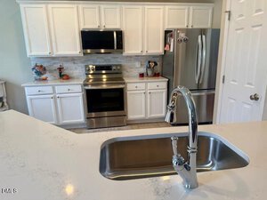 Modern kitchen with white cabinets, stainless steel appliances including a stove, microwave, and refrigerator, a marble countertop with a built-in sink and faucet in the foreground.