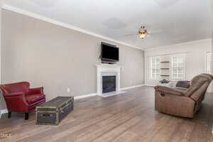 A living room with wood flooring, a beige wall, a white fireplace with a mounted TV, a brown recliner, a red armchair, a black trunk used as a coffee table, and windows with white blinds.