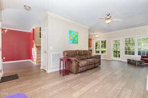 A spacious living room with light wood floors, beige walls, a brown sofa, small red side table, ceiling fan, colorful floral painting, and large windows showing greenery outside. Stairs with red accent wall are visible.