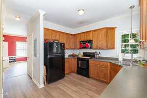 A kitchen with wooden cabinets, black appliances, a double sink under a window, and gray countertops. There is light wood flooring and a partial view of an adjacent red dining room.