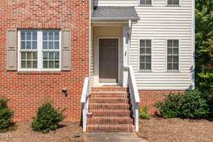 A brick and siding house exterior with a gray front door at the top of brick steps, white railings, two windows with white shutters, and green bushes along the foundation.