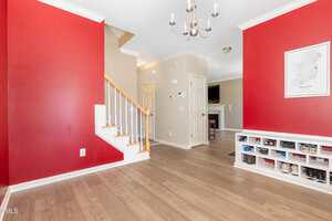 A room with red and beige walls, wooden flooring, a staircase with white railings, a shoe cubby, a chandelier, and a wall map of Ireland; a living area with a TV is visible in the background.