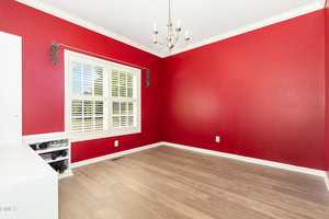 A room with bright red walls, white trim, a large window with white shutters, a small white built-in shelf with shoes, a light wood floor, and a simple chandelier hanging from the ceiling.
