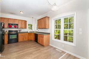 A kitchen with wooden cabinets, black appliances, and laminate wood flooring. There is a large window next to the sink and another large window in the dining area, letting in natural light.