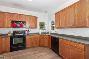 A kitchen with wooden cabinets, black appliances including a stove and dishwasher, gray countertops, two small windows above the sink, and light wood flooring. Red oven mitts hang on the stove.
