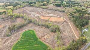 Aerial view of a large, partially cleared area of land with patches of remaining trees, a dirt road winding through, and some farmland adjacent to a paved road and houses in the lower right corner.