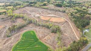 Aerial view of a large, mostly cleared area of land with some patches of trees, a winding dirt road, a small green field, and a few scattered houses near the edges.