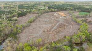 A large, cleared plot of land with visible dirt paths and minimal vegetation, surrounded by trees and forested areas; residential buildings are visible in the background.