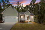 A modern, light gray single-story house at 407 Watson Street in Creedmoor features a double garage, white trim, and a front porch. Surrounded by trees at dusk with a colorful sky, two orange pumpkins sit by the front steps.