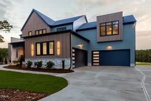 Modern two-story house with blue and brown exterior, large windows, and stone accents. The driveway is wide and curves toward the attached three-car garage. The yard has fresh mulch and neatly trimmed grass.