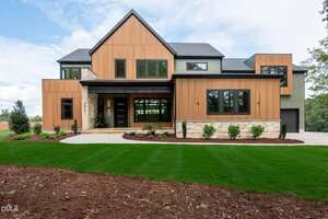 A modern two-story house at 3841 Graham Sherron Road in Wake Forest, featuring wood and stone exterior accents, large windows, a covered entryway, and a landscaped front yard with green grass and shrubbery.