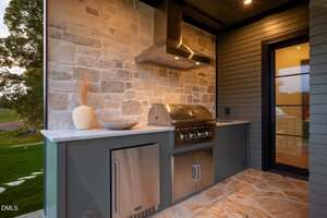 An outdoor kitchen with a stainless steel grill, range hood, mini fridge, and cabinets set against a stone wall. A decorative vase and shallow bowl sit on the countertop. Stone tile flooring is visible.