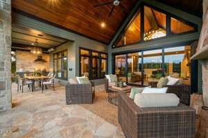 A covered outdoor patio features wicker furniture with light cushions, a dining table with chairs, stone flooring, and large windows and glass doors leading into a modern home with wood-paneled ceilings and warm lighting.