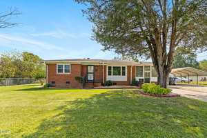 A single-story brick house with white trim, a covered front porch, landscaped bushes, a large tree in the front yard, and a driveway with a carport on the right side. The lawn is well-maintained.