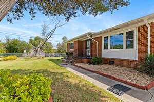 Single-story red brick house with white trim, a front porch, and a neatly landscaped yard with grass, shrubs, and a gravel-lined garden bed. A chain-link fence and trees are visible in the background.