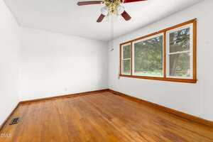 Empty room with wood flooring, white walls, large window with wood trim, and a ceiling fan with lights. The room is well-lit with natural light coming through the window.