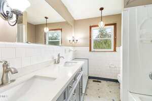 A bathroom with a double sink vanity, two mirrors, light fixtures, white tile walls, two windows, a toilet, and a shower with a curtain on the right. The floor has a light-colored pattern.