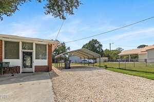 A gravel driveway leads to a double carport next to a brick house with large windows. A chain-link fence runs along the right side, enclosing a grassy yard. Trees and a neighboring house are visible in the background.