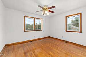 An empty room with white walls, two windows with wooden trim, hardwood floors, and a ceiling fan with lights. The room is well-lit with natural light coming through the windows.