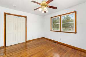 Empty room with polished hardwood floors, white walls, two windows with wooden trim, a ceiling fan with lights, and a closed double-door closet. Natural light enters through the windows.