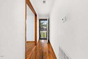 A narrow hallway with white walls, a wooden floor, a thermostat on the right wall, and a metal security door at the end leading outside. An open doorway on the left leads to another room with wood flooring.