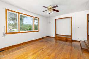 Empty room with hardwood floors, white walls, large windows, a ceiling fan with lights, and a recessed area with two steps leading up. Daylight comes through the windows, and there is no furniture present.