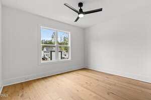 A bright, empty room with light wood flooring, white walls, a large window showing houses and trees outside, and a black ceiling fan with a light in the center of the ceiling.